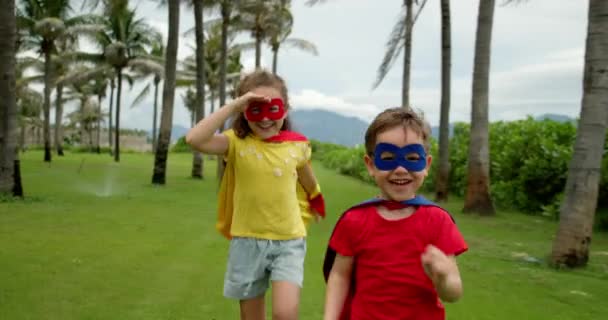 Retrato duas crianças engraçadas bonitos, menino e menina vestidos com trajes em super-heróis, em capas vermelhas e máscaras azul-vermelhas, de mãos dadas, olhar para os olhos uns dos outros, em seguida, correr para a frente demonstra seu poder herói — Vídeo de Stock