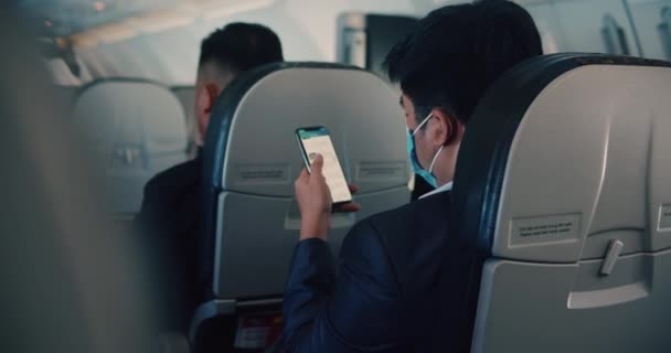 En affärsman med asiatiskt utseende flyger på ett flygplan, använder en telefon på ett flygplan, bläddrar igenom bilder, meddelanden, öppnar en ansökan. — Stockvideo