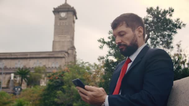 Schwerer bärtiger Mann im teuren Anzug tippt etwas auf seinem Handy, während er draußen auf einer Parkbank sitzt — Stockvideo