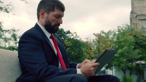 Homme d'affaires mature lisant un journal assis dans le parc sur le banc. Hommes d'affaires avec journal et tablette assis sur un banc dans un parc à l'extérieur. — Video