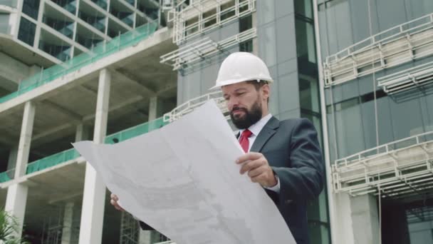 Mühendis, beyaz kasklı bir erkek inşaat alanında çok katlı binaların çizimlerini kontrol ediyor.. — Stok video