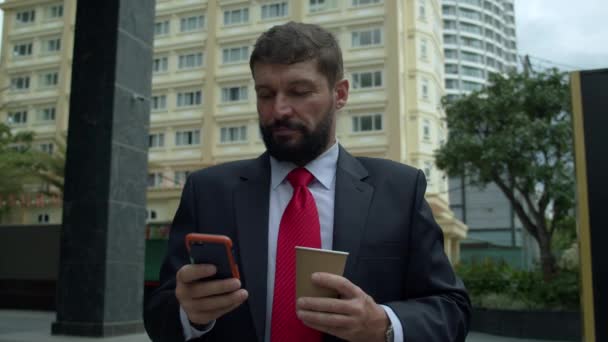 Homme barbu élégant sérieux dans des vêtements astylish textos sur smartphone personnel, la lecture d'un journal sur une tablette en plein air. Homme d'affaires avec téléphone, boire du café. — Video