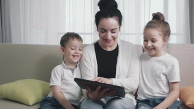 Mutlu bir anne ve tatlı bir kız çocuğu, anne evde çocukla oynuyor rahatlamak için akıllı bir telefon kullanıyor kanepeye oturuyor, komik videolar izliyor, eğleniyor, aile hayatının tadını çıkarıyor.