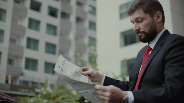Geschäftsmann senior sitzt im teuren Anzug mit schönem Bart und schwarzblauer Jacke auf einer Bank im Park und liest Zeitung. — Stockvideo
