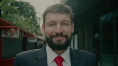 Pahalı takım elbiseli, yakışıklı, sakallı kıdemli bir işadamı sokakta binaların yanında kameraya bakar..