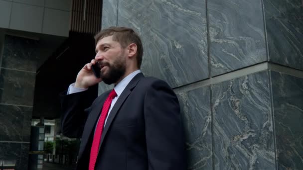 Un homme parle sur un portable dans la rue. Un homme d'affaires sérieux d'âge moyen en vêtements luxueux dans le quartier financier d'une métropole utilise un téléphone portable pour les négociations. 4K — Video