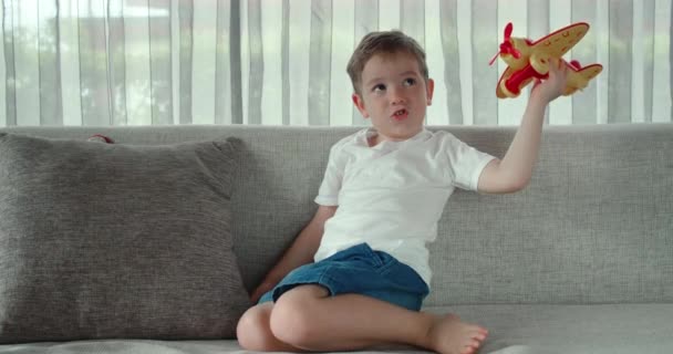 Un simpatico bambino in età prescolare che gioca a casa sul divano con un aeroplano giocattolo, un simpatico bambino seduto sul divano lancia un aeroplano nelle sue mani. — Video Stock