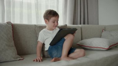 Çocuklar teknoloji bağımlılığı konsepti. Aletli bir çocuk. Evdeki koltukta oturmuş dijital tablet teknolojisi kullanan sevimli bir anaokulu çocuğu..