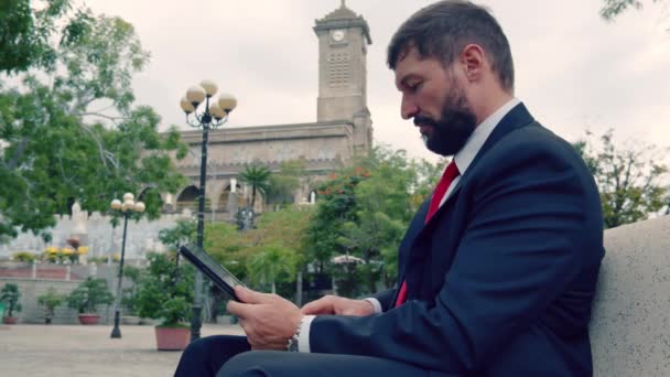 Homme d'affaires mature lisant un journal assis dans le parc sur le banc. Hommes d'affaires avec journal et tablette assis sur un banc dans un parc à l'extérieur. — Video
