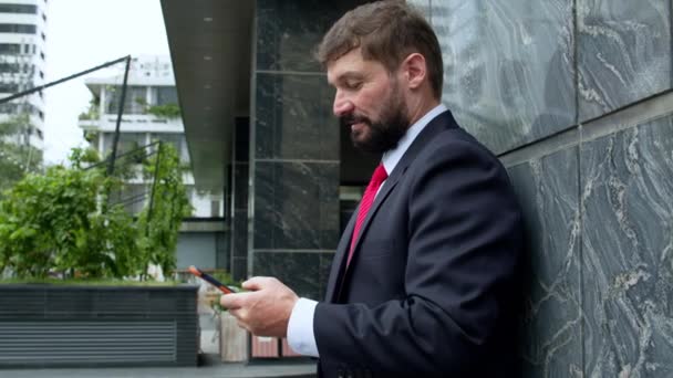 Un homme d'affaires envoie un texto dans la rue. Un homme d'affaires sérieux d'âge moyen en vêtements luxueux dans le quartier financier d'une métropole utilise un téléphone portable pour les négociations. 4 — Video