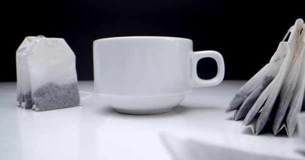 Super primer plano conjunto de bolsas de té en una mesa blanca sobre un fondo negro y una taza de té blanco.Hacer té de la mañana.Este clip de té es adecuado para proyectos culinarios y culinarios. — Vídeo de stock