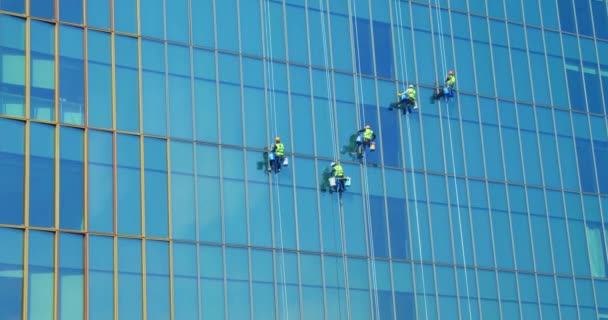 Öt védő kovás maszkos munkás, kék és sárga munkaruhában mossa egy üzleti felhőkarcoló külső ablakait - az ipari hegymászást. A magas emberek ablakot mosnak. Tisztítási koncepció — Stock videók