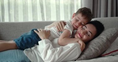 Mutlu bir anne ve oğul, kanepede birlikte dinleniyorlar, bu anaokulu öğrencileri annelerinin karnında uyuyorlar, evde sarılıyorlar, anne çocuğun kafasını okşuyor..