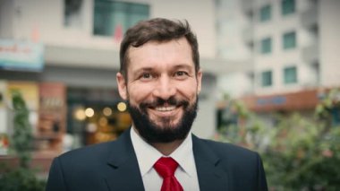 Pahalı takım elbiseli, yakışıklı, sakallı kıdemli bir işadamı sokakta binaların yanında kameraya bakar..