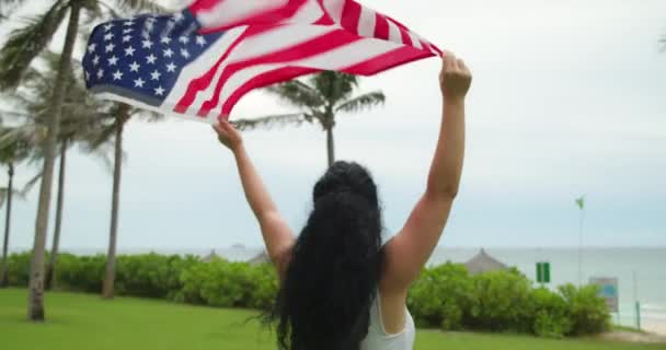 El concepto de patriotismo, día de la independencia y días festivos - una joven feliz sonriente con una camiseta corre con la bandera nacional americana en el verano en el parque. — Vídeo de stock