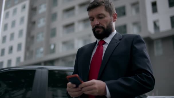 Un homme d'affaires envoie un texto dans la rue. Un homme d'affaires sérieux d'âge moyen en vêtements luxueux dans le quartier financier d'une métropole utilise un téléphone portable pour les négociations. — Video