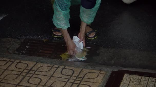 A cleaning oldman on the street removes garbage. Cleanliness and order in the yard depends on the janitor during quarantine.Rainwater flows on the road and fall into the metal hatch with holes drain. — Stockvideo