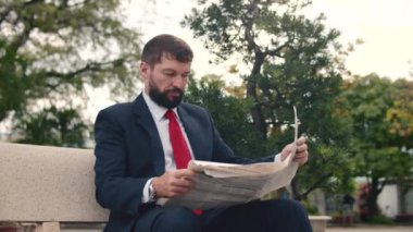 Soft focus footage Businessman senior in an expensive suit with a beautiful beard in black blue jacket sits on bench in the park and reads a newspaper.
