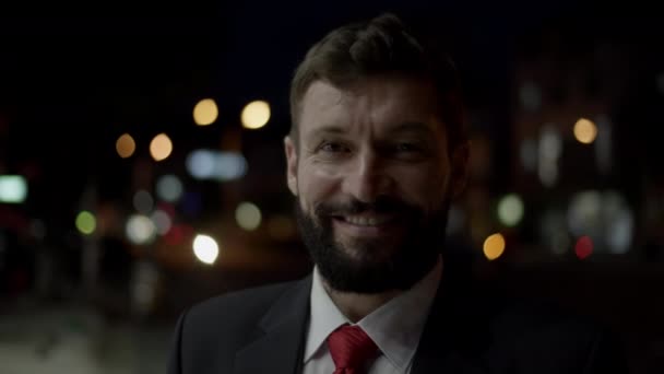 Портрет чоловіка в дорогому костюмі, красивий бородатий старший бізнесмен ввечері стоїть на вулицях міста поруч з бізнес-будівлею і дивиться на камеру, посміхаючись . — стокове відео