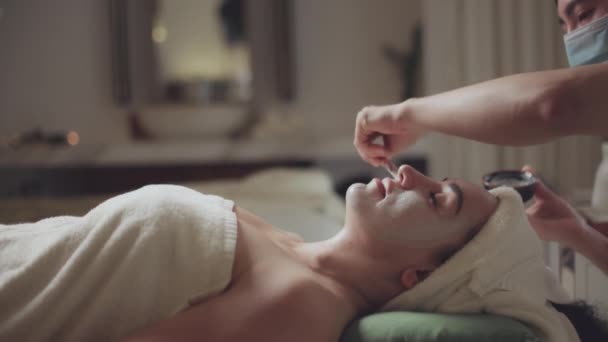 Relaxovaná žena ležící na lázeňském lůžku pro lázeňskou léčbu masáže obličeje a hlavy masérem v luxusním lázeňském středisku, dívka s obličejovou maskou v křečích. — Stock video