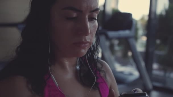 Portrait a young active sportswoman engaged in a woman in the gym does exercise on a stationary bike, listens to music while doing sports. — Stockvideo