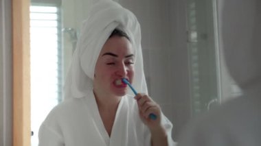 Oral Bakım Konsepti. Beyaz dişli, beyaz dişli beyaz kadın, evde izole edilmiş aynaya bakıyor. Banyoda dişlerini fırçalayan ve gülümseyen sevimli genç kız. Yaşam biçimi.