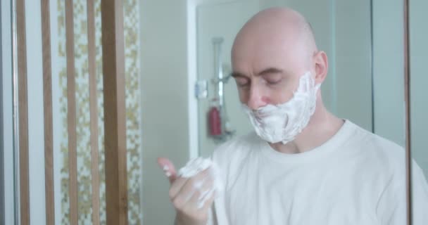 Среднего возраста бритье с использованием одноразовой бритвы в ванной комнате, крупным планом, вид сбоку. Мужчина наносит крем для бритья на лицо, гигиеническая процедура. Человек бреет бороду, часть лица в кадре. — стоковое видео