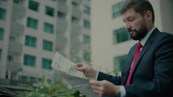 Senior middle-aged in an expensive suit with a beautiful beard in black blue jacket sits on bench in the park and reads a newspaper. — Stockvideo