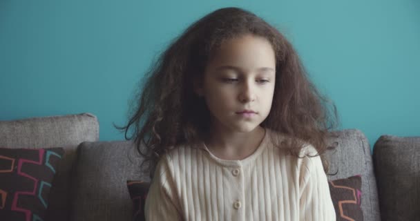 Portrait of a sad little girl, an upset child lowering her eyes to the floor and then looking at camera, sits on the sofa at home,a cute child with a beautiful face sits on sofa. Sad childhood concept — Stok Video