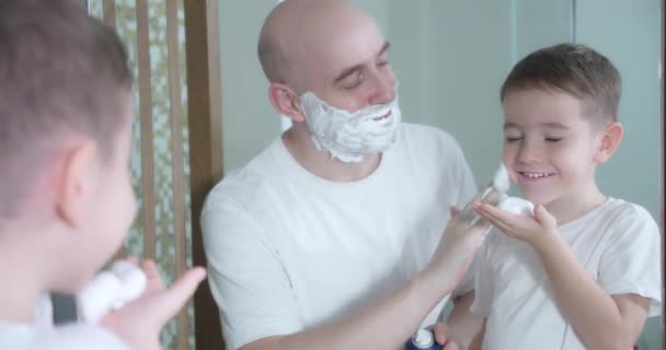 Padre enseña a su hijo a afeitarse. Hijo pequeño entretiene al padre se aplica crema de afeitar en la cara. Padre e hijo juegan con crema de afeitar en la cara. El hombre aplica crema de afeitar a la cara — Vídeos de Stock