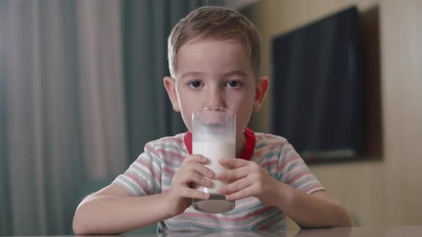 Retrato de criança caucasiana feliz menino feliz que bebe leite de um copo enquanto está sentado na cozinha em casa. Conceito de infância feliz gostando de passar o tempo em casa. — Vídeo de Stock