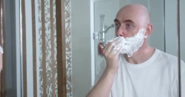 Orta yaşlı, banyoda tek kullanımlık tıraş bıçağı kullanıyor. Yakın plan, yan görüş. Adam tıraş kremini yüz yüze sürüyor, hijyenik prosedür. Adam sakalını tıraş ediyor, yüzünün bir parçası çerçevede..
