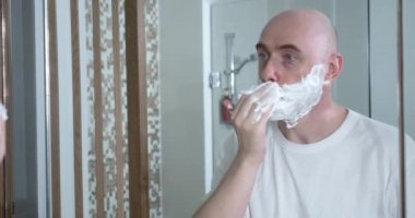 Orta yaşlı, banyoda tek kullanımlık tıraş bıçağı kullanıyor. Yakın plan, yan görüş. Adam tıraş kremini yüz yüze sürüyor, hijyenik prosedür. Adam sakalını tıraş ediyor, yüzünün bir parçası çerçevede..