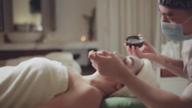 Sağlık, stres atma ve gençleştirme konsepti. Spa yatağında uzanıp spa tedavisi gören rahat bir kadın. Spa merkezindeki masör, spa merkezinde yüz maskesi takan bir kız..