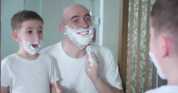 Father teaches son to shave. Little son entertains the father applies shaving cream to his face. Father and child play with shaving cream on their face. Man applies shaving cream to face — Stock Video