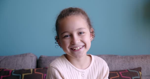 카메라를 보면서 웃고 있는 이상 한 어린 소녀가 소파에 앉아 있습니다. 귀엽고 얼굴 이 예쁜 아이가 소파에 앉아 있습니다. 행복 한 어린 시절에 대한 개념. — 비디오