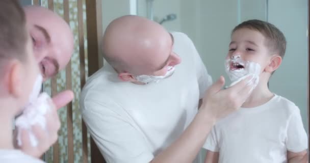 Padre enseña a su hijo a afeitarse. Hijo pequeño entretiene al padre se aplica crema de afeitar en la cara. Padre e hijo juegan con crema de afeitar en la cara. El hombre aplica crema de afeitar a la cara — Vídeos de Stock