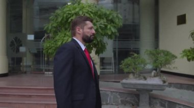 Pahalı takım elbiseli kıdemli işadamı elinde evrak çantasıyla güneşli yaz gününde şehir caddesinde ağır çekimde yürüyor. Sokaklarda yürüyen bir iş adamı.