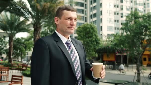 The shot moves around a businessman standing on street in the city center, who and drinks hot coffee,sun shine,enjoying beautiful morning feeling happy.Portrait of corporate businessman smiling — Vídeo de Stock