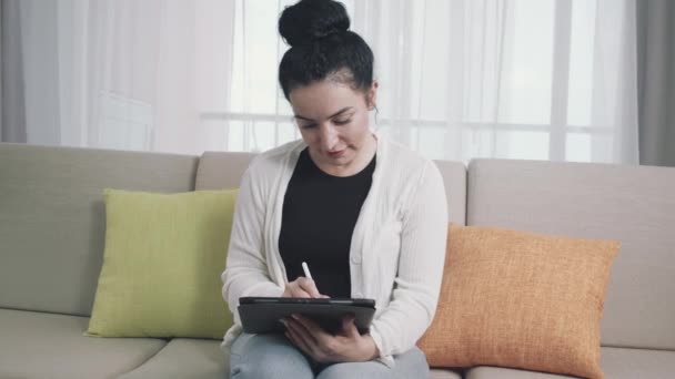 Žena sedí doma na pohovce, kreslí, píše a používá pero s digitálním tabletovým počítačem. Projev bystré, veselé energie jako tvůrce, tvůrce něčeho nového, profesionálního — Stock video