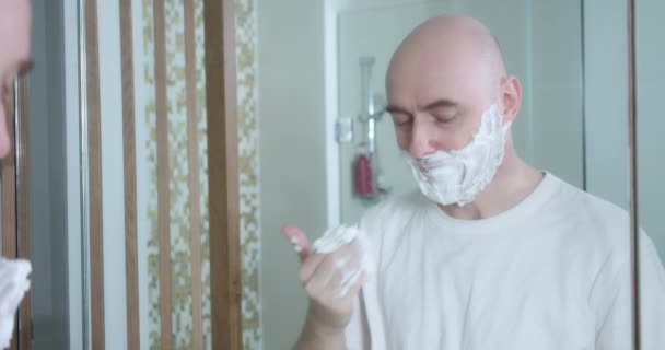 Среднего возраста бритье с использованием одноразовой бритвы в ванной комнате, крупным планом, вид сбоку. Мужчина наносит крем для бритья на лицо, гигиеническая процедура. Человек бреет бороду, часть лица в кадре. — стоковое видео