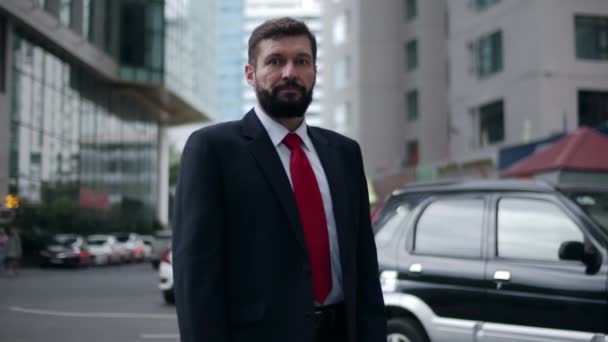 Homem de negócios em um terno caro e uma gravata vermelha, um belo empresário de barba sênior à noite fica nas ruas da cidade ao lado de um prédio de negócios e olha para a câmera, sorrindo — Vídeo de Stock
