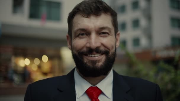 Портрет бізнесмена в дорогому костюмі і червоній краватці, красивий бородатий старший бізнесмен ввечері стоїть на вулицях міста поруч з бізнес-будівлею і дивиться в камеру, посміхаючись — стокове відео