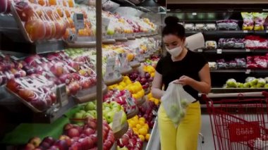 Coronavirüs salgınından maske takan genç bir kadın bir süpermarketin market bölümünde duruyor. Meyve alıyor, yeşil elma..