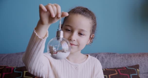 Porträt Lustiges kleines Mädchen lächelndes Kind, das einen Spielzeugball anschaut, sitzt zu Hause auf der Couch, niedliches Kind mit hübschem Gesicht, das Ball mit einer Überraschung betrachtet. Konzept glückliche Kindheit. — Stockvideo