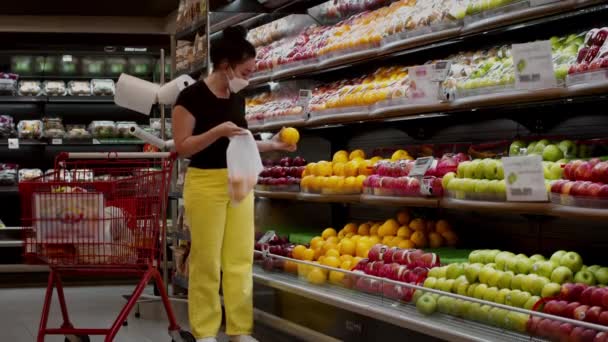 Een jonge vrouw met een masker van de coronavirusepidemie staat in de kruidenierswinkel van een supermarkt sinaasappels in een zak te stoppen. Ze koopt fruit, groene appels.. — Stockvideo