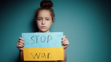 Ukraynalı zavallı çocuk korkmuş evsiz kız savaş çatışmasını protesto ediyor. Afişi yazı ile kaldırıyor. Mavi arka planda savaş yok. Barış, saldırganlığı bırak, çocuk Rus savaşına karşı.