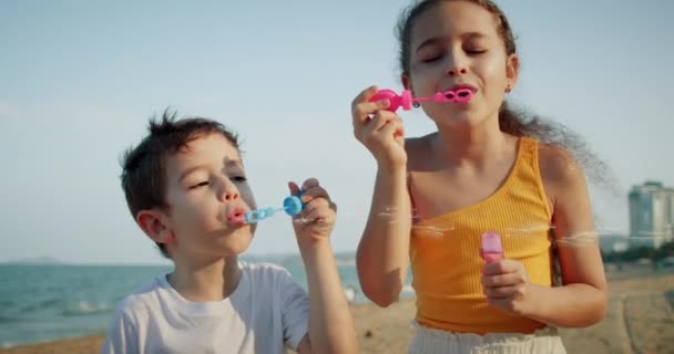 Szczęśliwe dzieci bawiące się bańkami mydlanymi.Dzieci szczęśliwy uśmiech pęcherzyki wydmuchu na plaży. Portret uśmiecha się jak małe, kaukaskie dzieci. Brat i siostra pękają bańki mydlane. Gry Dzieci zabawy na świeżym powietrzu gry — Wideo stockowe