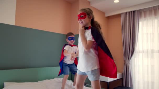 Zwei Kinder im rot-blauen Superman-Kostüm, Mädchen und Jungen, springen im Zimmer auf dem Bett, im Kinderzimmer.Superhelden, Bruder und Schwester, spielen zu Hause und stellen sich vor, sie seien Superhelden. — Stockvideo