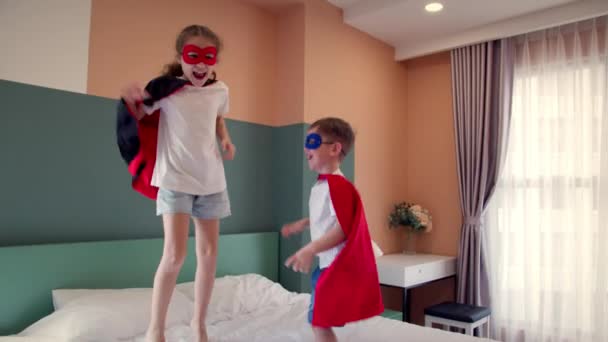 Két gyerek piros és kék Superman jelmezben, lány és fiú szuperhősök, ugrálnak a szobában az ágyon, a gyerekszobában.Szuperhősök, testvér és testvér, játszanak otthon képzelve, hogy ők szuperhősök. — Stock videók